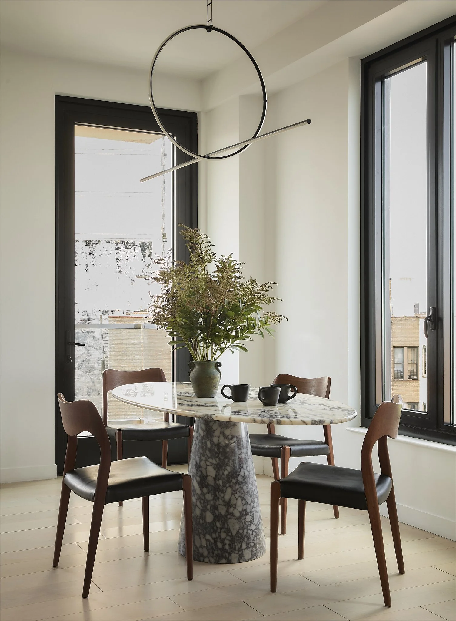 Prospect Heights dining area by Hallie Rose Design featuring a round, dramatically veined marble pedestal table, vintage wood-and-leather chairs, sculptural circular pendant lighting, and black-framed windows.