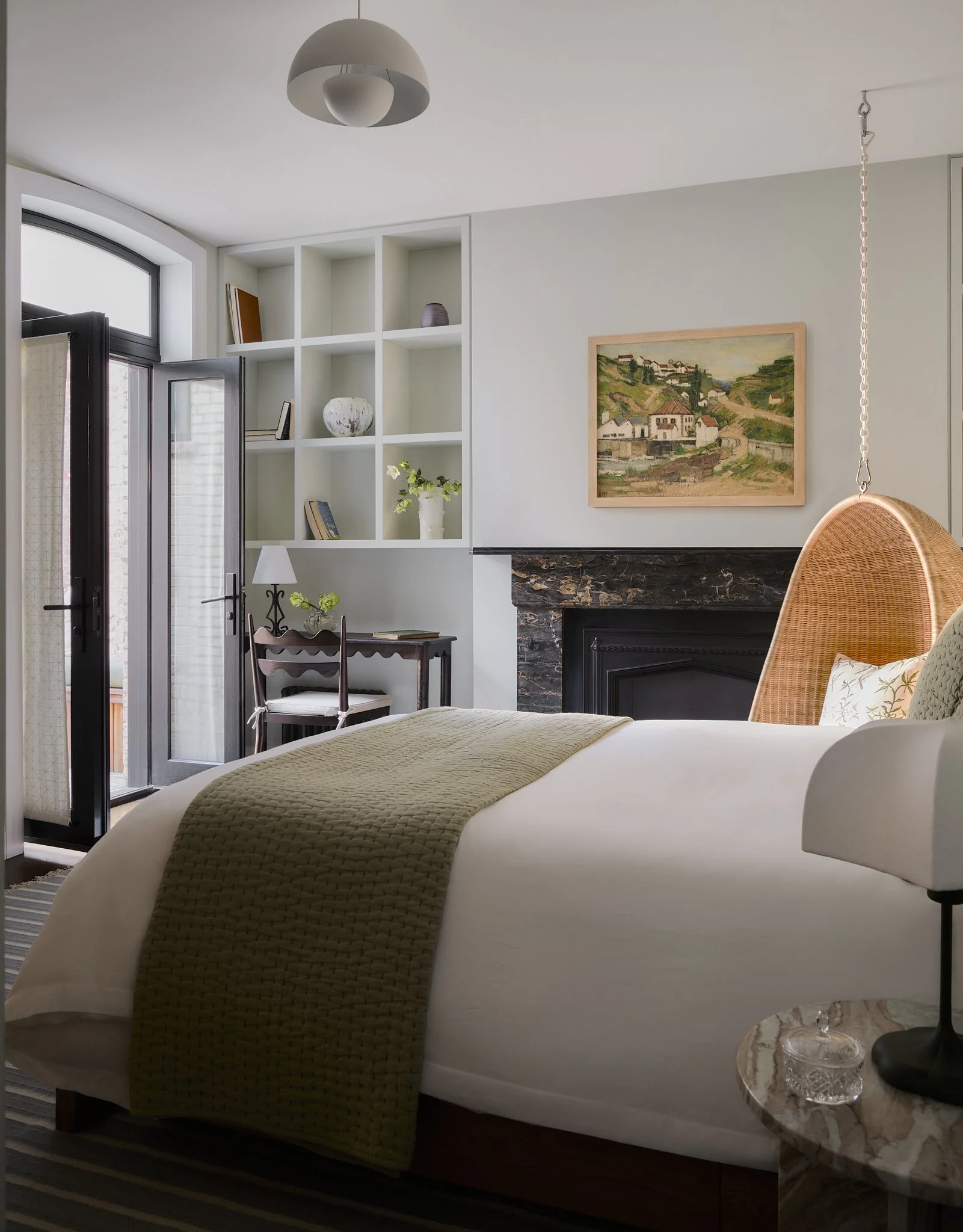 Brooklyn Heights primary bedroom by Hallie Rose Design featuring a suspended rattan chair, black marble fireplace, built-in shelving, vintage writing desk, and olive-green textiles.