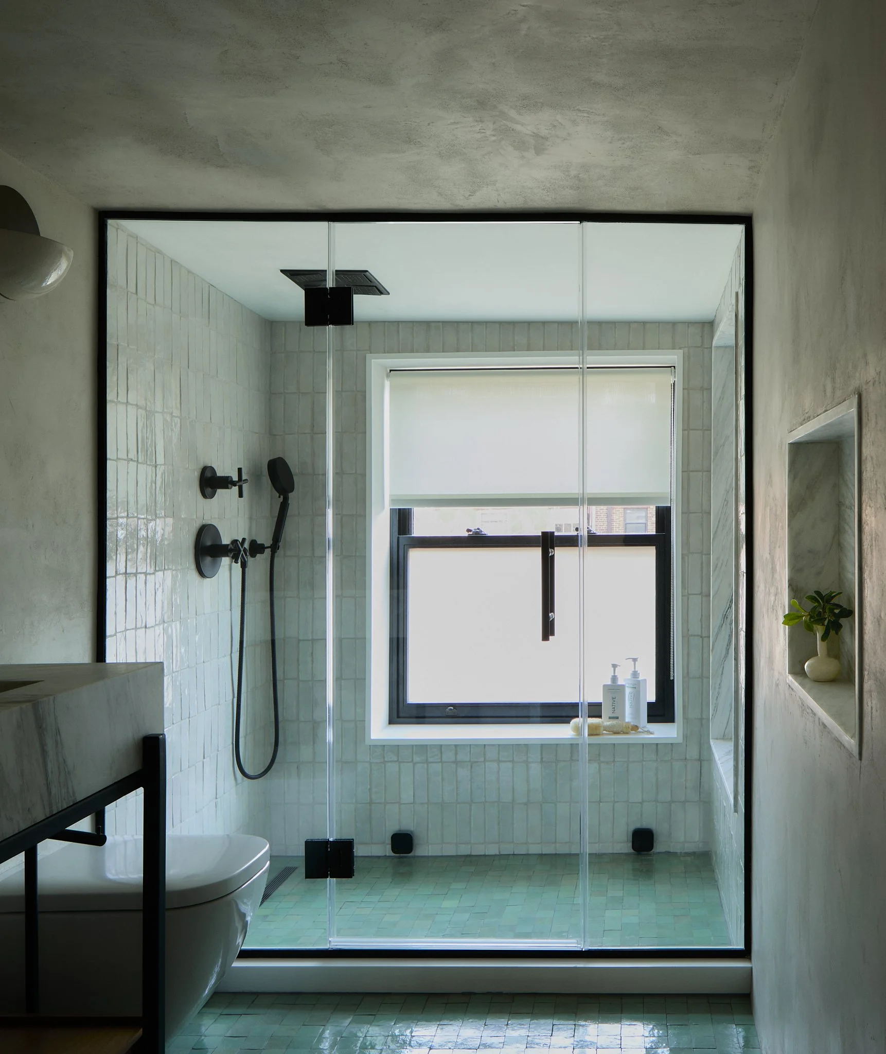 Brooklyn Heights bathroom by Hallie Rose Design featuring a frameless walk-in shower, pale green mosaic floor tile, handmade-look glazed wall tile, stone niches, and matte-black fixtures.