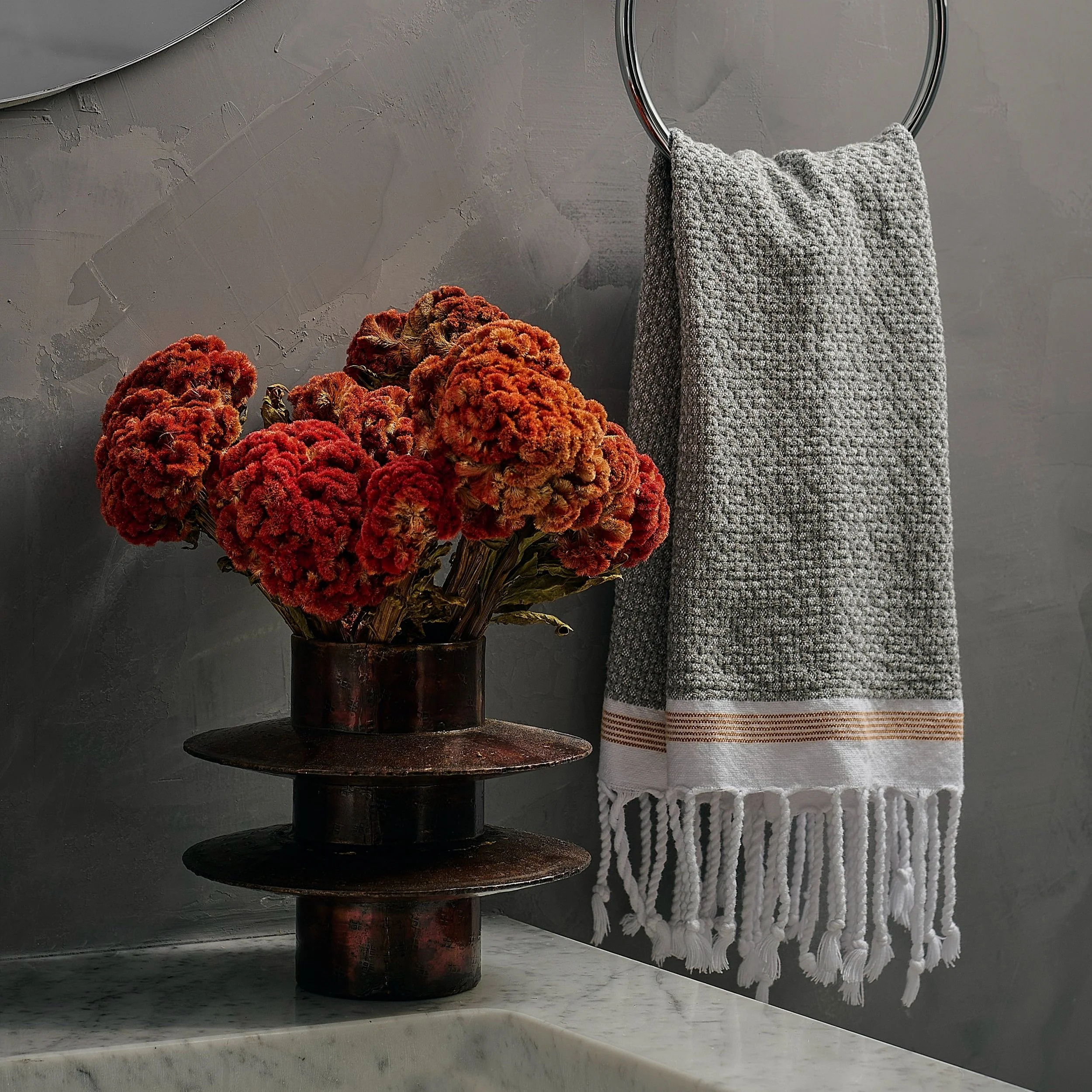 Textural guest bathroom detail styled by Hallie Rose Design with red celosia flowers, a sculptural dark metal vessel, and a fringed woven textile.