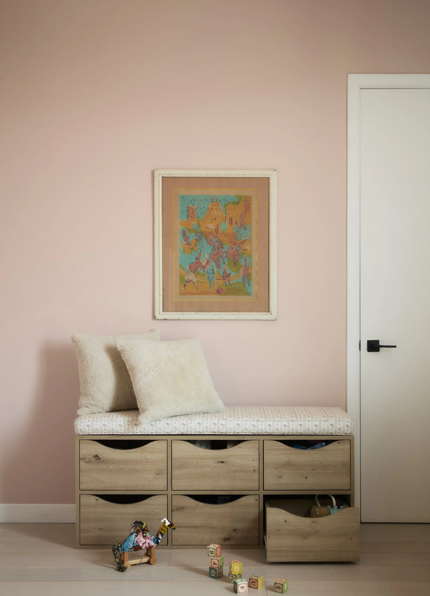 Prospect Heights children’s room by Hallie Rose Design featuring an oak toy-storage bench, floral upholstered cushion, bouclé pillows, blush-pink walls, vintage artwork, and wooden toys.