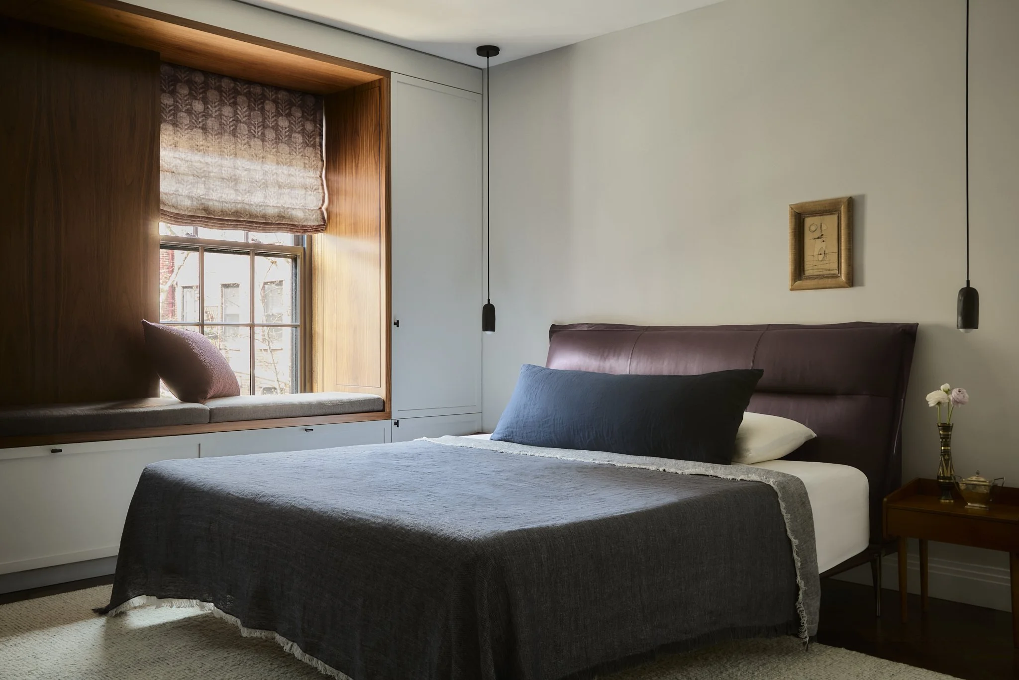 A cozy bedroom with a large bed dressed in a dark gray bedspread and pillows. There is a window seat with a pink pillow, a window with wooden trim, and a roman shade. A framed artwork hangs above the bed, and black pendant lights are suspended on either side. A bedside table with a vase of flowers is visible, and beige carpet covers the floor.