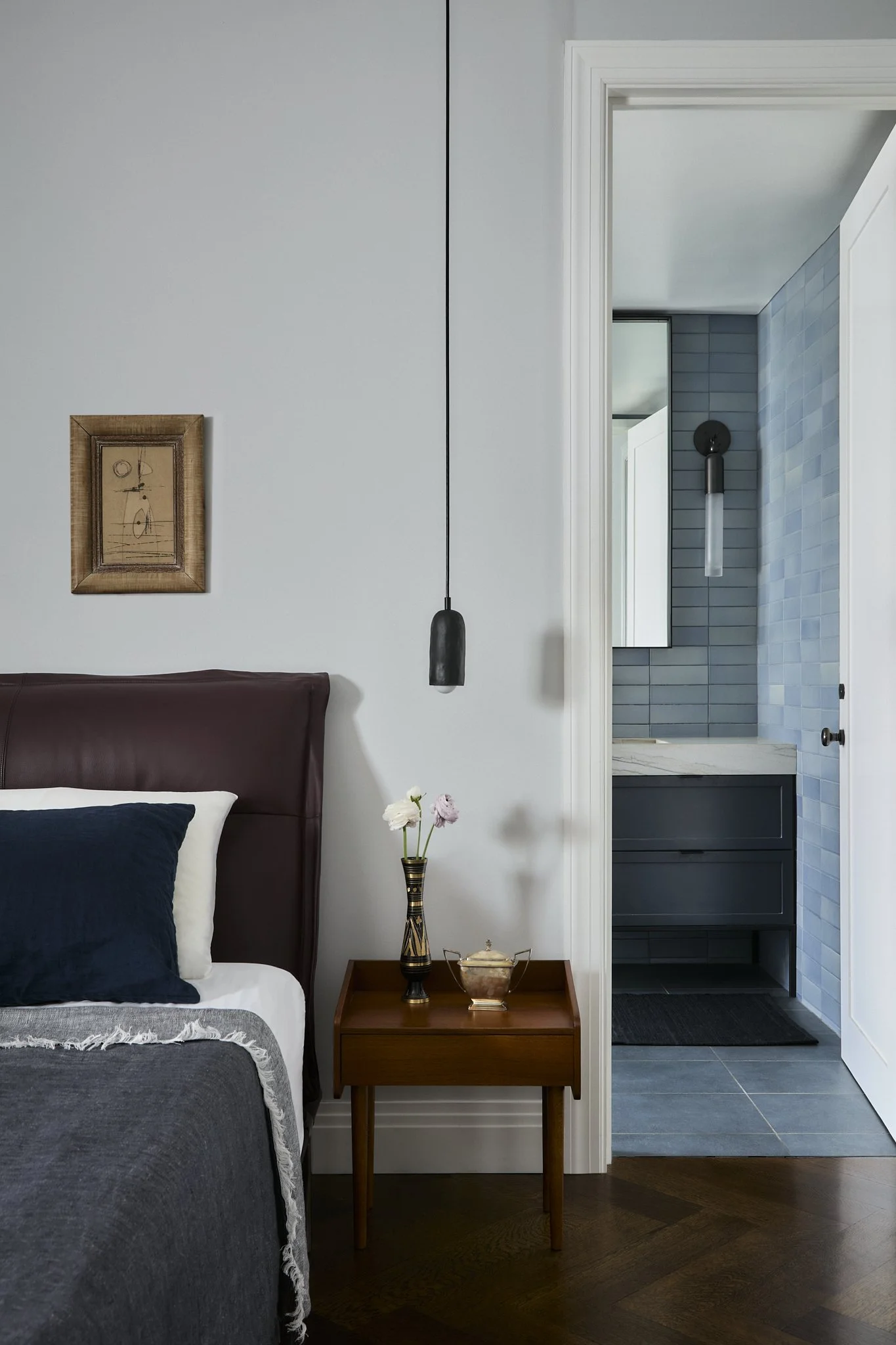 Brooklyn Heights primary bedroom by Hallie Rose Design featuring a burgundy leather headboard, vintage wood nightstand, sculptural pendant light, and blue ceramic-tiled ensuite bathroom.