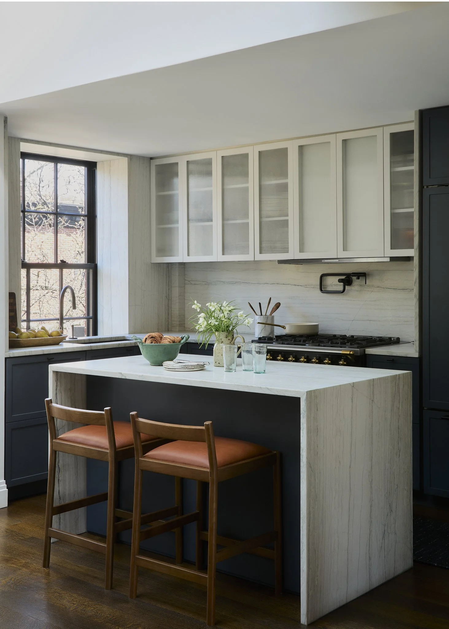 Modern kitchen with white upper cabinets, a large window, a kitchen island with a marble countertop, two wooden chairs with orange cushions, and various kitchen items on the counter.
