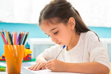 Young girl sitting at a desk, focused on her work, with colored pencils in a cup nearby.