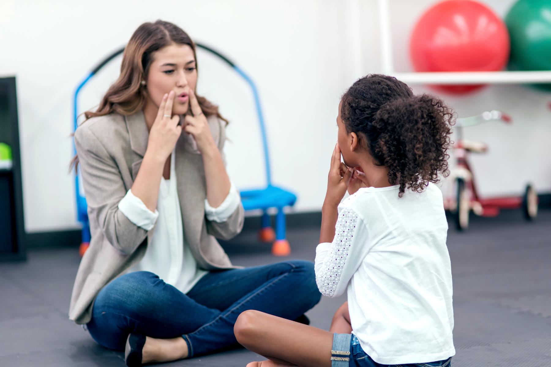 A woman and a girl sitting cross-legged on the floor facing each other, both with fingers pointing to their faces, appearing to practice facial exercises or expressions, inside a room with therapy or exercise equipment.