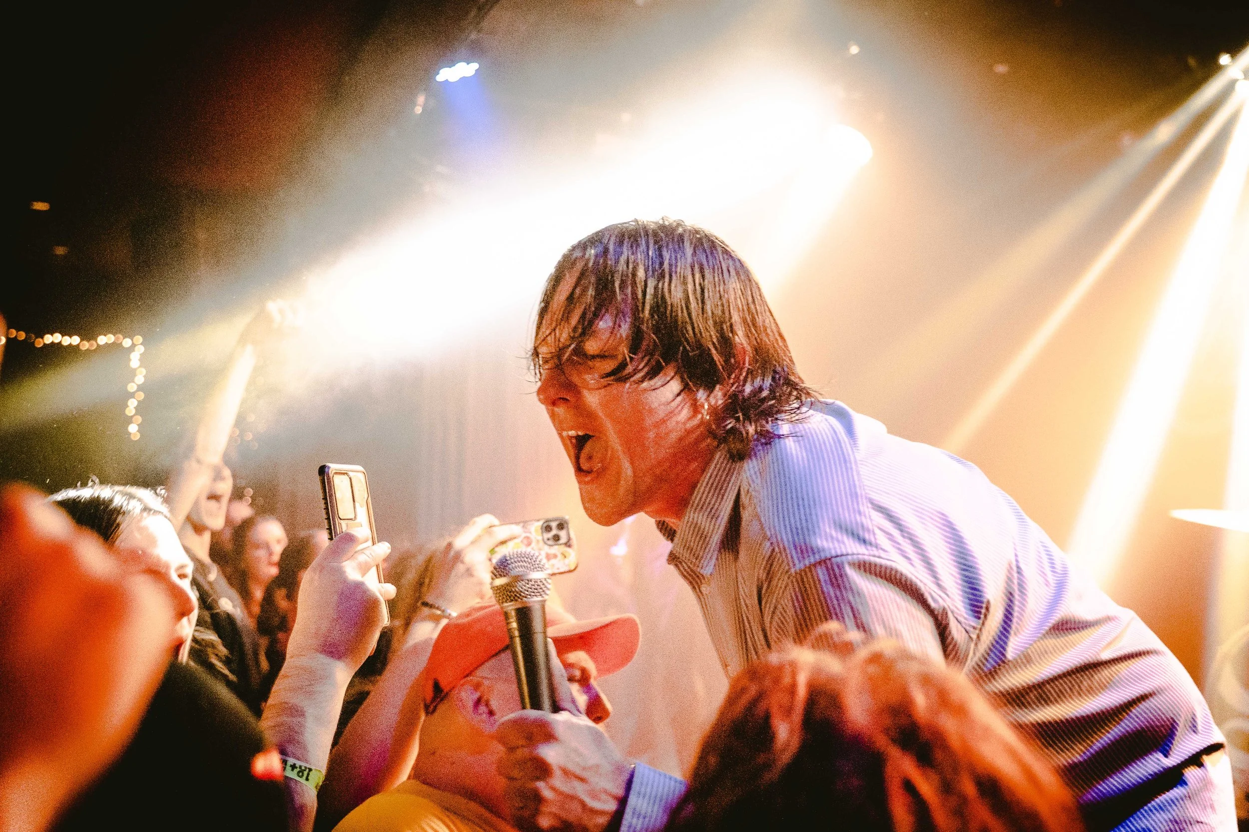 Photograph of a male performer singing passionately into a microphone at a live concert, surrounded by audience members taking photos.