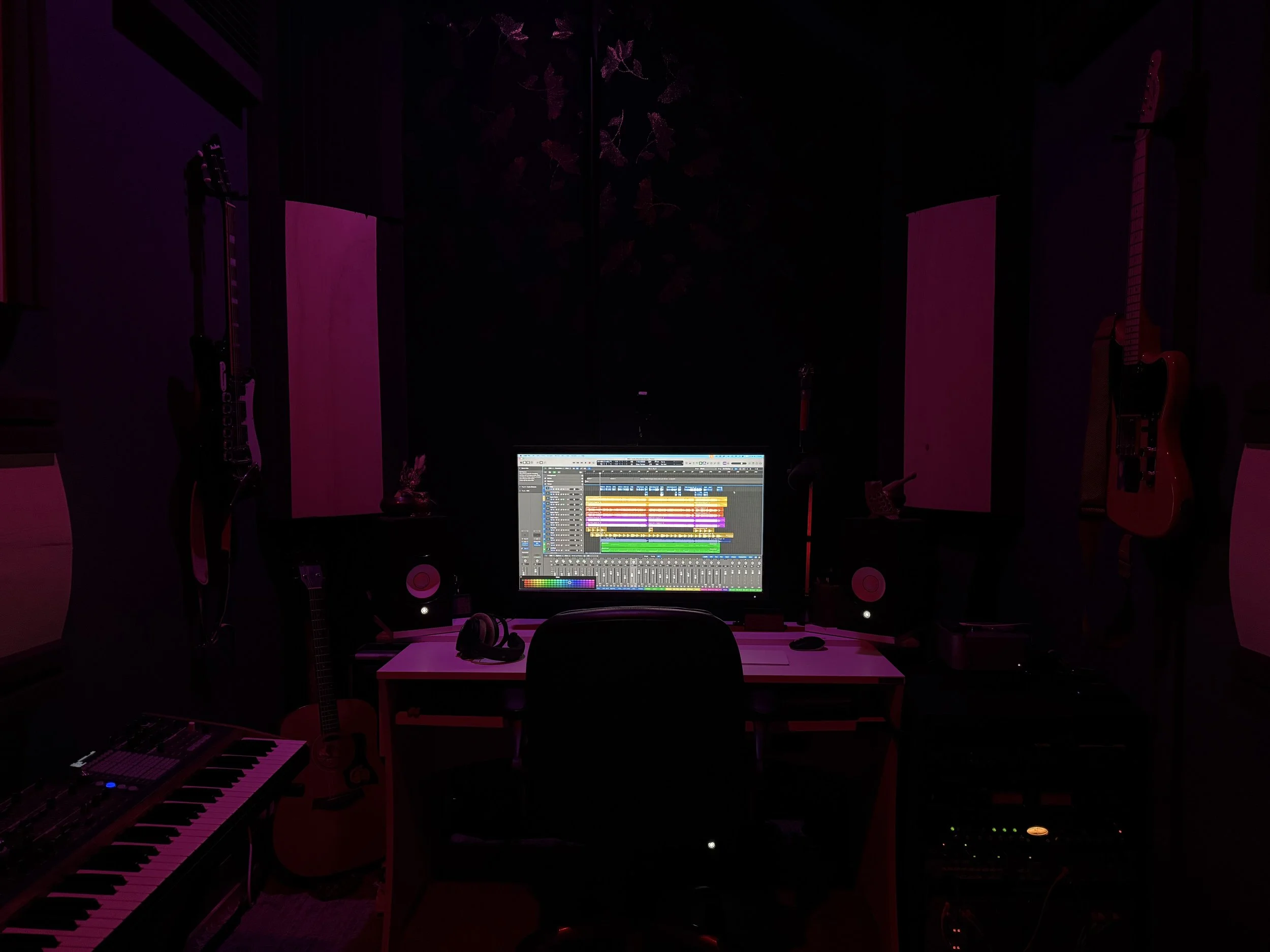 Music studio with a computer displaying digital audio workstation, surrounded by guitars, speakers, keyboard, and headphones, illuminated by pink and purple lighting.