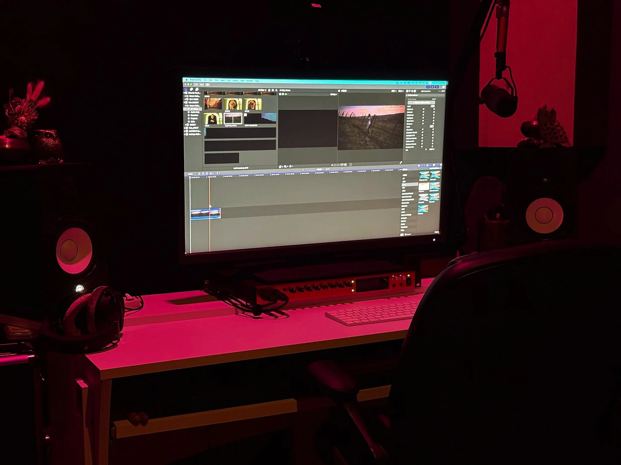 Computer workstation with a large monitor displaying video editing software, surrounded by studio speakers, headphones, and decorative items in a dark room with red lighting.