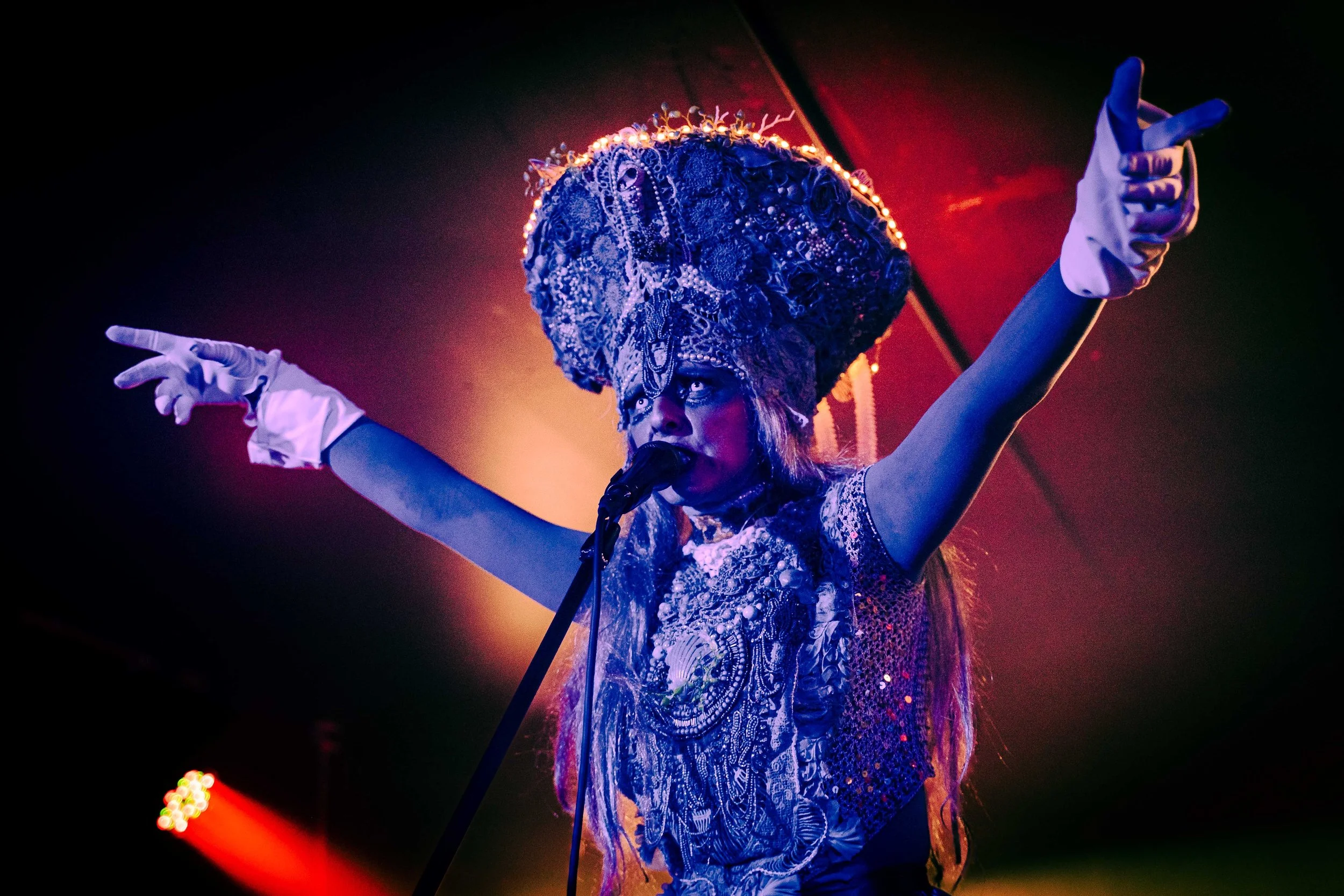 Performer wearing an elaborate headpiece and ornate dress, singing into a microphone with arms outstretched on stage, colorful lighting background.