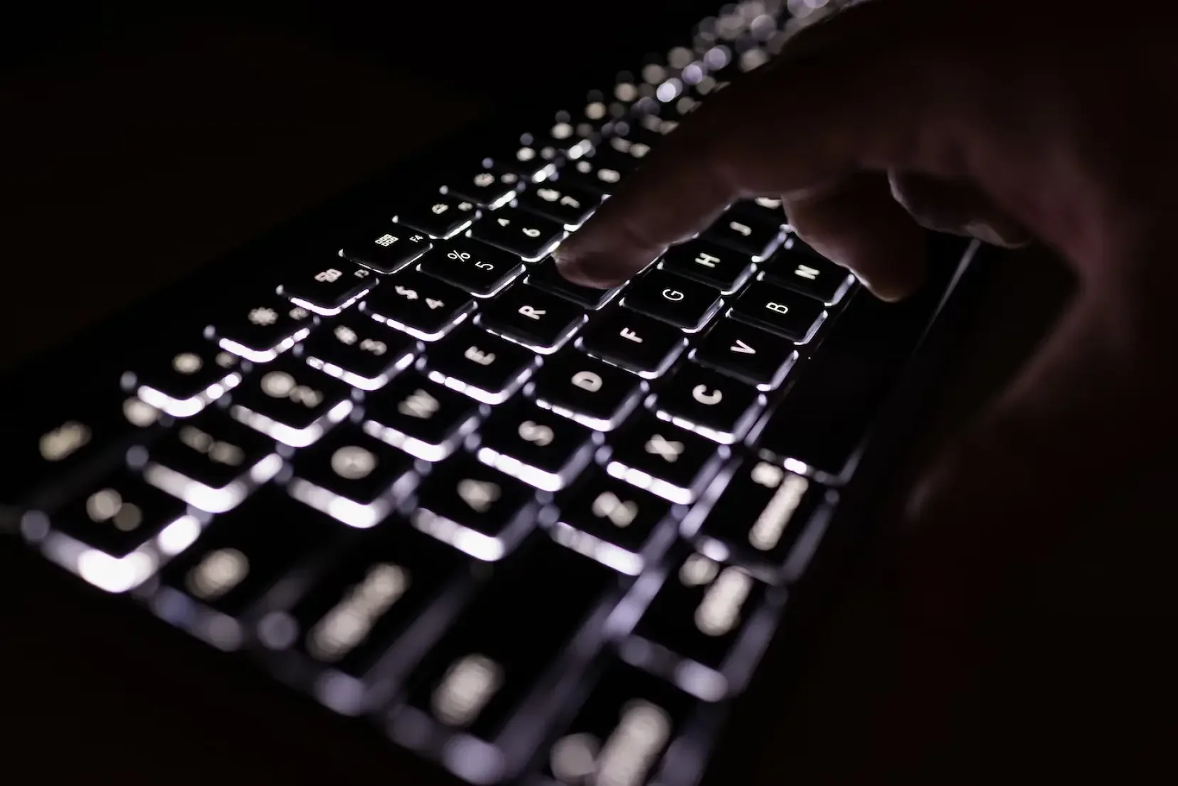 A person's hand typing on a backlit computer keyboard in a dark environment.