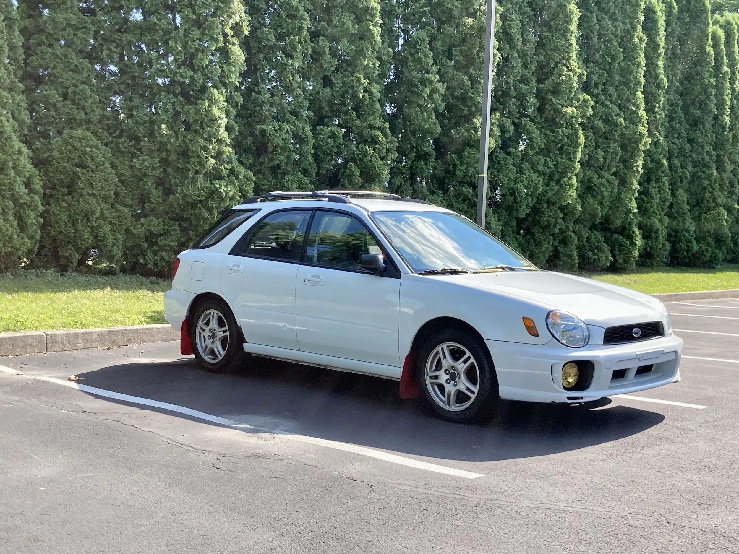 A white car parked in a parking lot with a tree line in the background.