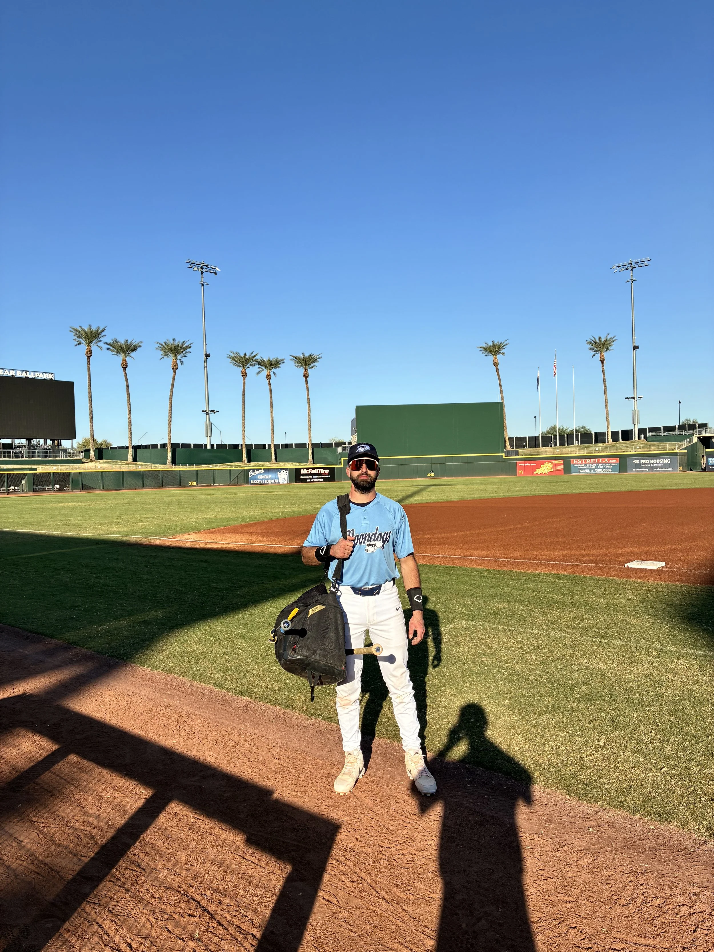 A man in a light blue sports jersey, white pants, and white sneakers stands on a baseball field holding a backpack and a skateboard. The background shows a baseball outfield wall, tall palm trees, and a bright blue sky.