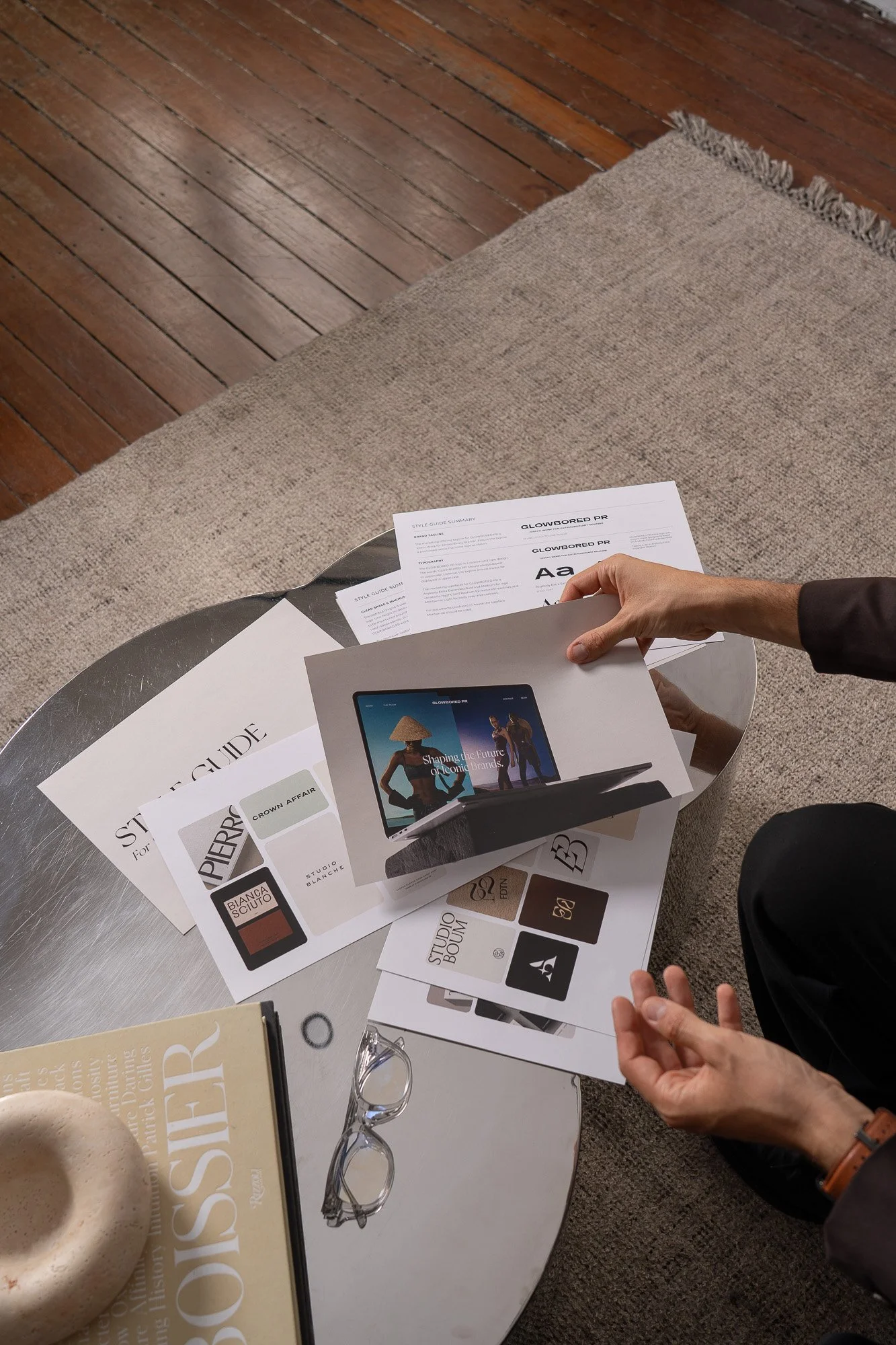 A person holding a page with the text 'GLOWBORED PR' over a table with brochures, color samples, a pair of glasses, and a book. The table is end with a beige fringe rug and a wooden floor.