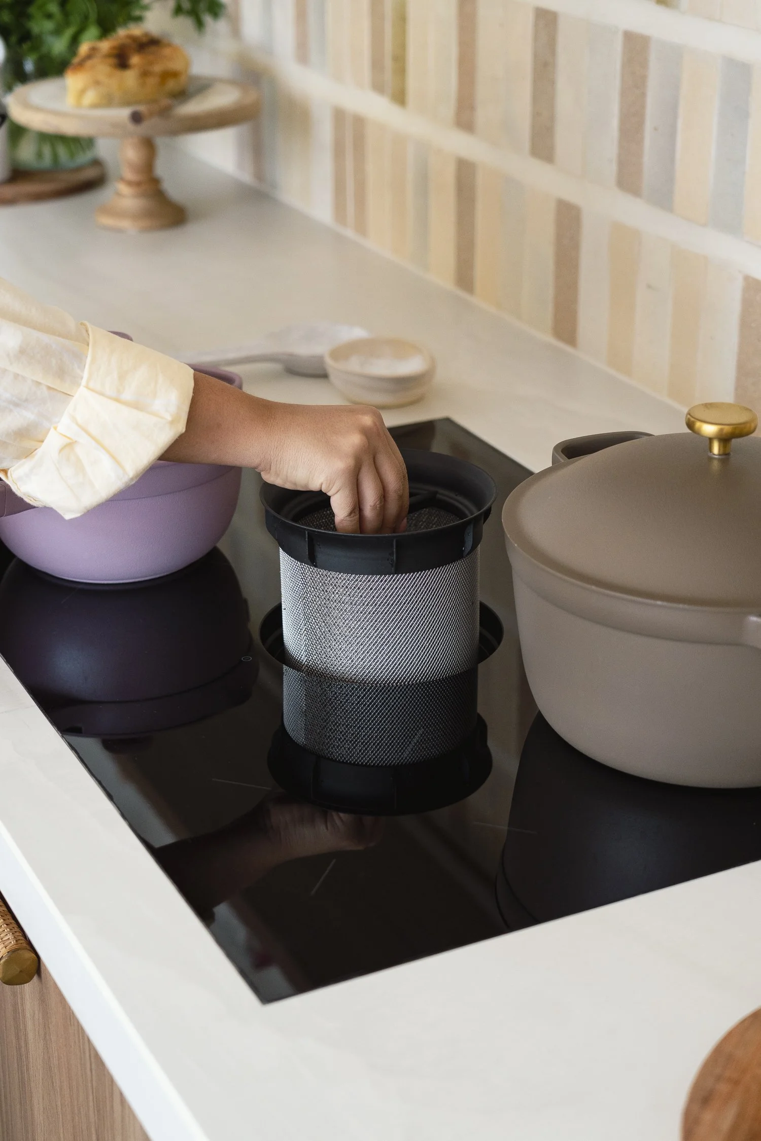 A person is reaching into a small mesh container on a black electric stovetop, with a purple mixing bowl and beige pot nearby, and some kitchen utensils and a pie on a wooden cake stand in the background.