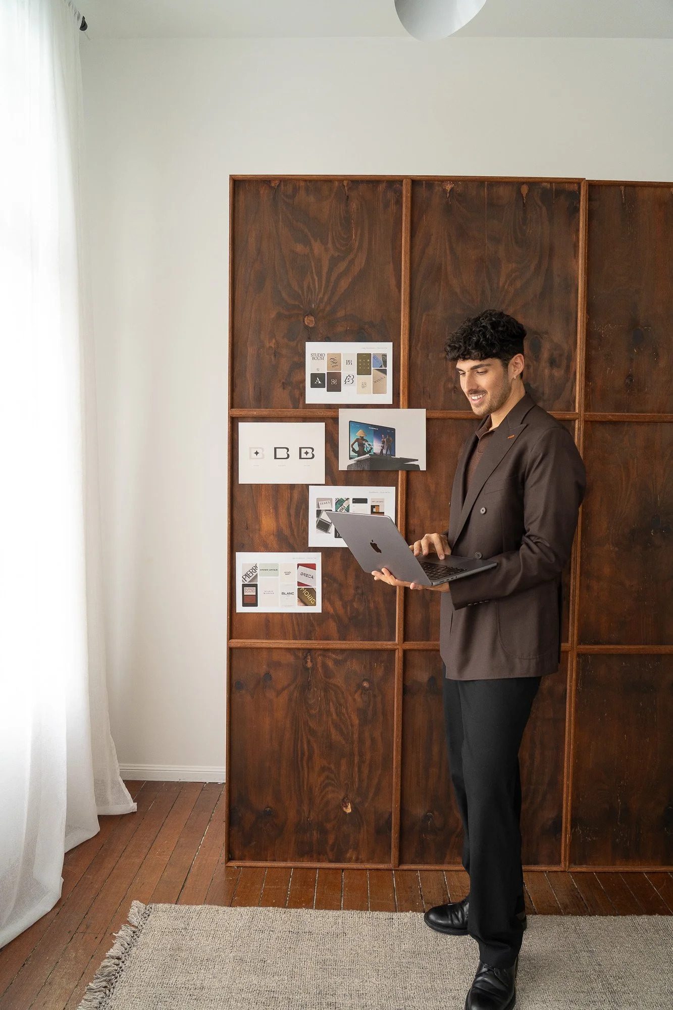 A man in a brown suit smiling and looking at a laptop in a room with a wooden partition wall and white curtains.