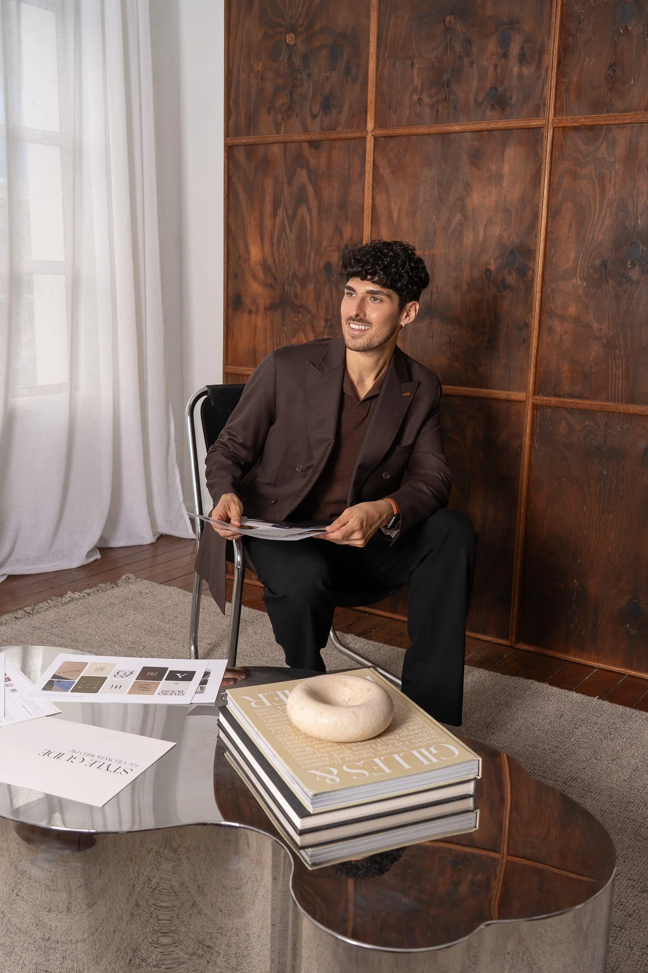 A young man with curly dark hair wearing a dark brown blazer and black pants is sitting in a modern black chair with a metal frame, holding a magazine, and smiling. He is positioned in front of a wooden wall panel. In front of him, there is a glass coffee table with magazines, papers, and a stone sculpture on top. The room has white curtains and a beige rug, with sunlight coming through the curtains.