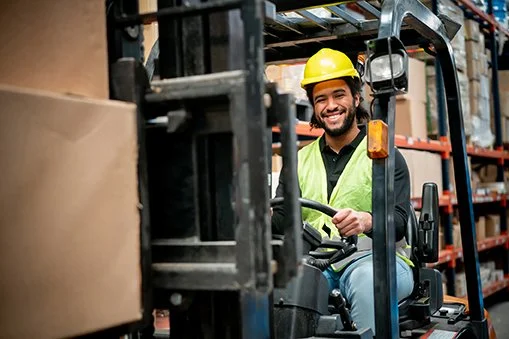 A man wearing a yellow safety helmet and a reflective safety vest smiling while operating a forklift in a warehouse.