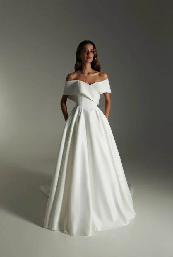 Ganzkörperaufnahme einer Frau in einem schlichten, eleganten Brautkleid mit schulterfreiem Carmen-Ausschnitt, tailliertem Oberteil und weit fallendem Rock; sie steht aufrecht vor hellem, neutralem Hintergrund.