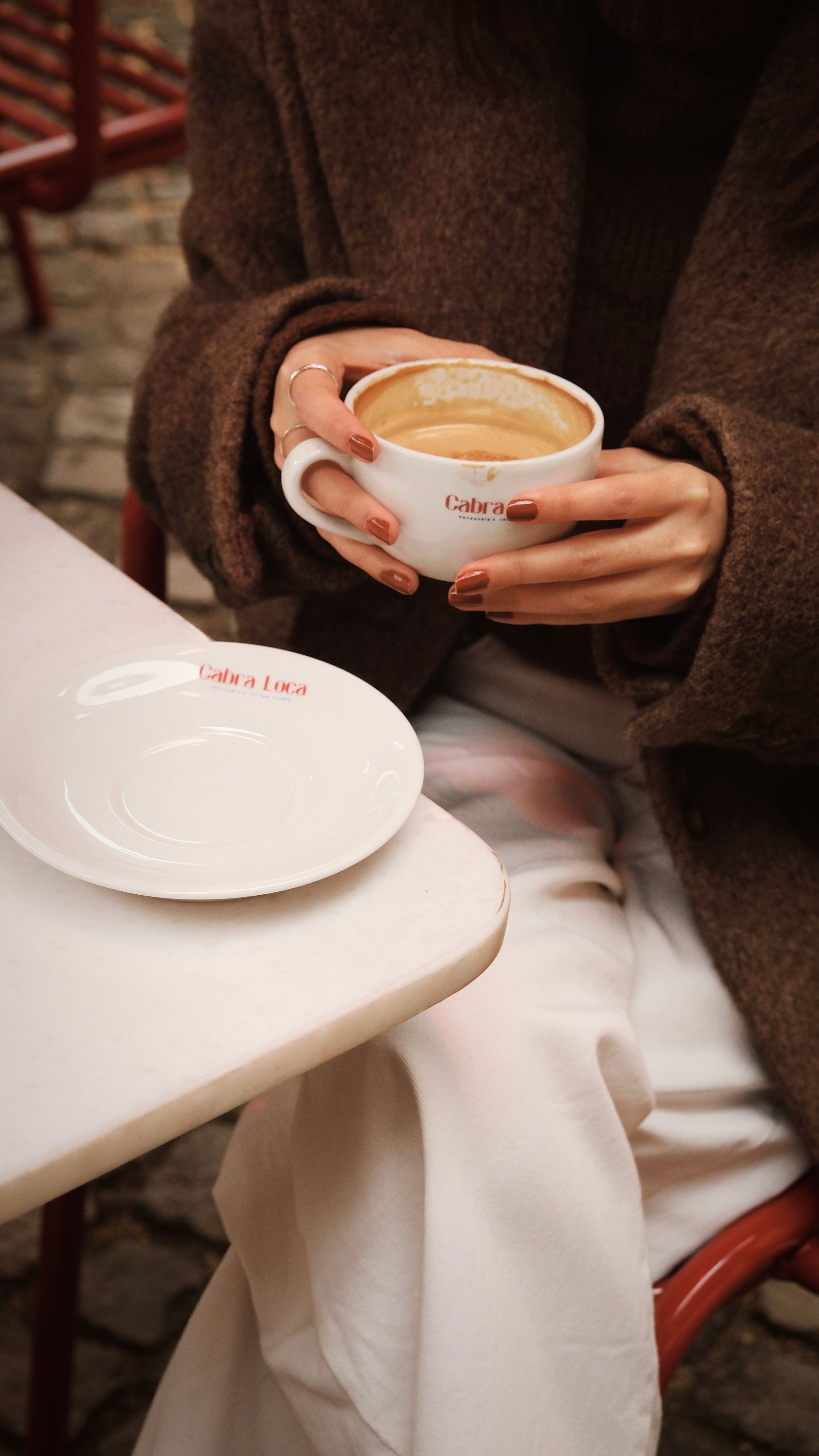Eine Person im braunen Mantel hält eine weiße Kaffeetasse an einem Café-Tisch, davor steht eine leere Untertasse mit „Cabra Loca“-Schriftzug.