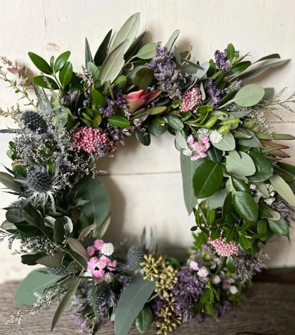 Nahaufgenommen: üppiger Blumenkranz an heller Wand aus Eukalyptus, Olive und Buchsbaum, verziert mit blauen Disteln, rosa-weißen Wachsblumen und lila Blütenständen.