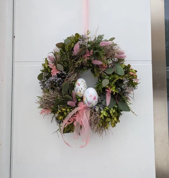 Festlicher Osterkranz an weißer Tür: grünes Reisig (Buchsbaum, Konifere), rosa Samtgras und kleine lila Blüten. Im unteren Drittel zwei weiße Ostereier mit floralem Schmetterlingsdruck, betont durch breite transparente rosa Schleife.