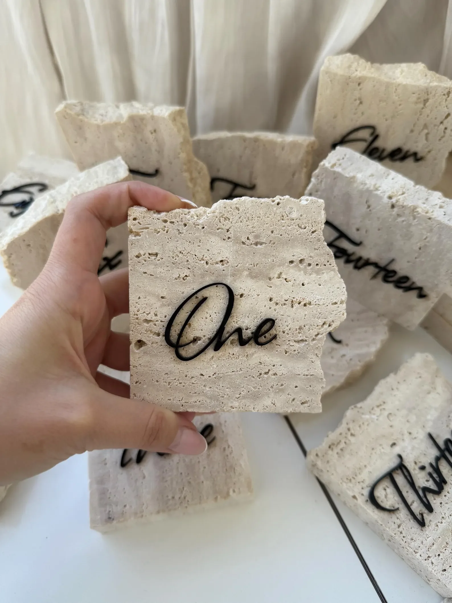 Eine Hand hält eine quadratische, raue Natursteinplatte mit der schwarzen Aufschrift „One“ in geschwungener Schreibschrift.