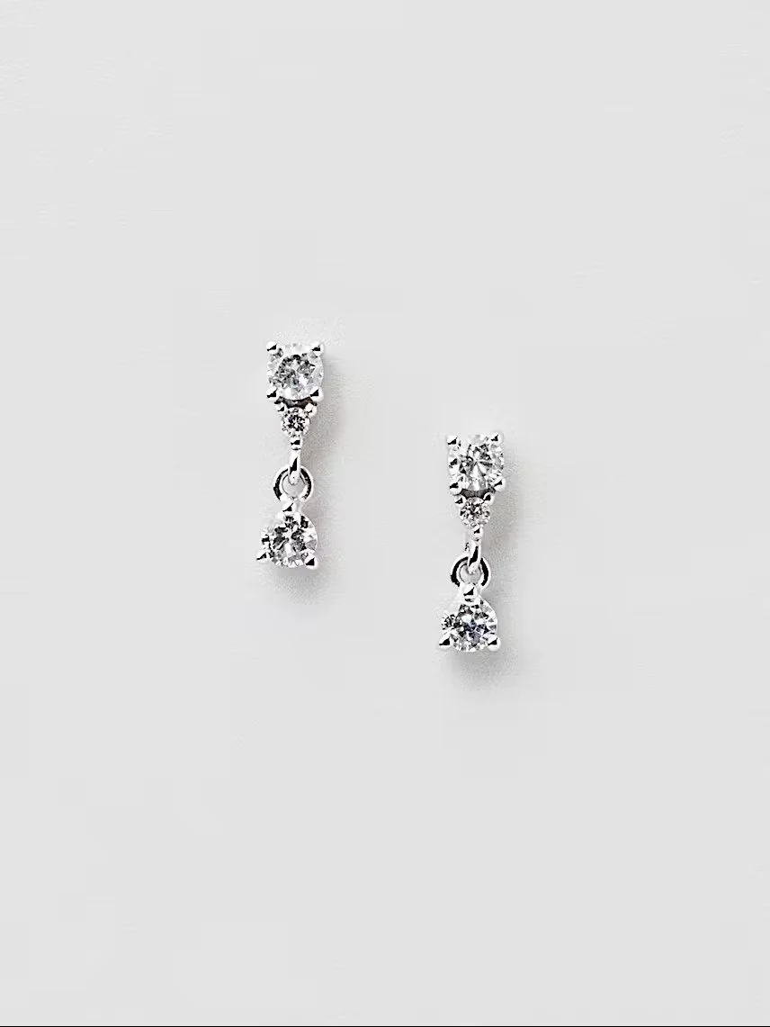 Paar hängende Ohrringe aus Silber vor hellem Hintergrund. Sie bestehen aus einem kleinen runden Stecker mit winzigem Kristall, an dem ein beweglicher, runder Glitzerstein hängt.