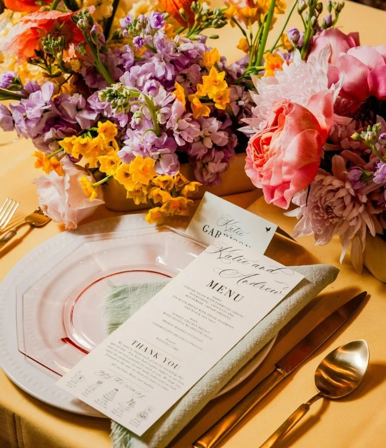 Ein gedeckter Tisch mit einem Blumenarrangement aus lilafarbenen, gelben und pinkfarbenen Blumen. Auf dem Tisch liegt ein weißer Teller mit einem Metallbesteck (Messer und Löffel), eine Besteckgabel, eine bedruckte Menükarte und eine Dankeskarte.