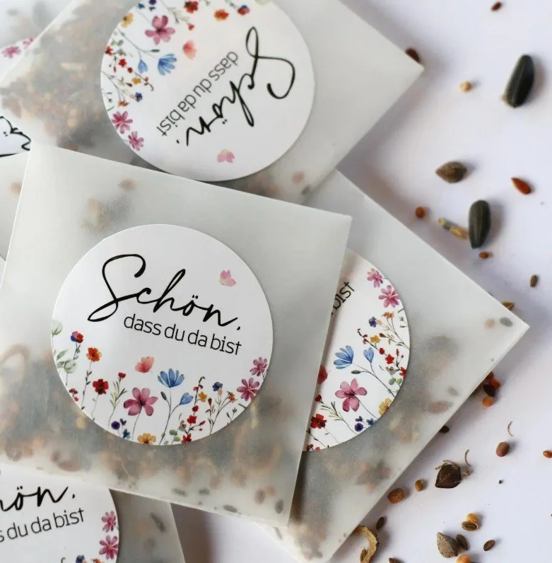 Kleine Tütchen mit Blumensamen als Gastgeschenk, verziert mit einem runden Aufkleber mit Blumenmuster und der Aufschrift „Schön, dass du da bist“.