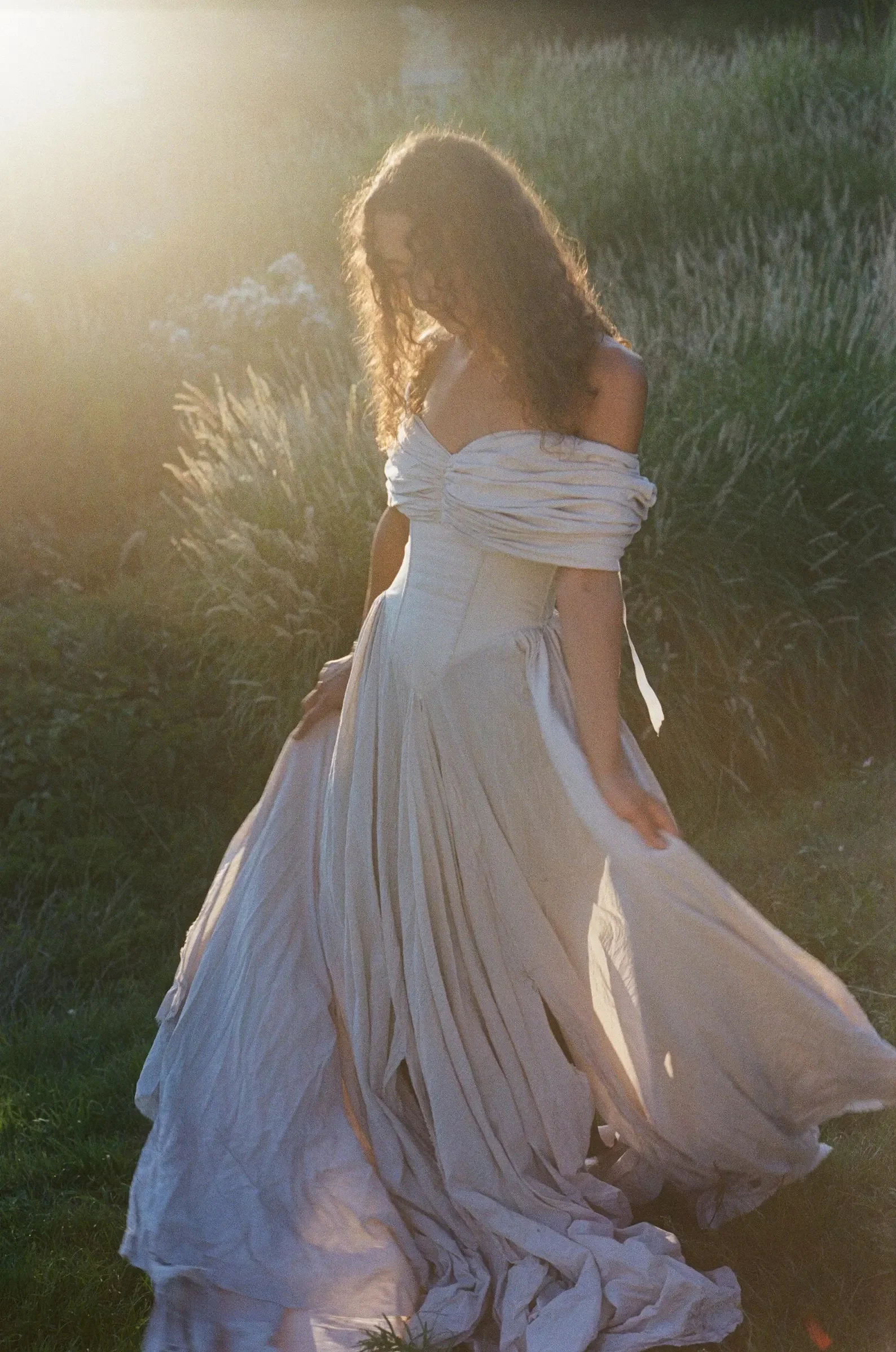 Braut in schulterfreiem Vintage-Kleid mit figurbetontem Korsett und weitem, fließendem Rock im hohen Gras, vor warmem, goldenem Gegenlicht im analogen Fotografie-Stil.