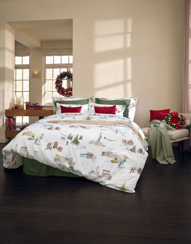 Weihnachtlich dekoriertes Schlafzimmer. Weißes Bett mit illustrierten Festtagsmotiven, roten und grünen Kissen und grünem Betthaupt, daneben ein Sessel mit Decke und Kranz.