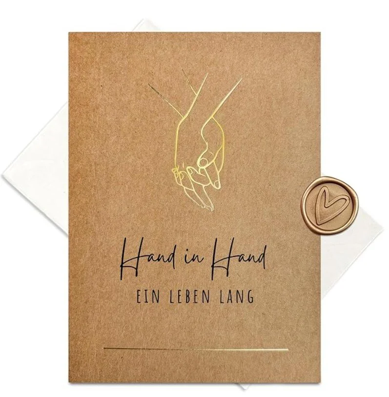 Elegante Glückwunschkarte zur Hochzeit aus braunem Kraftpapier. Das Design zeigt eine feine, goldene Linienzeichnung von zwei Händen, die sich halten, darunter der Text: „Hand in Hand – EIN LEBEN LANG“.