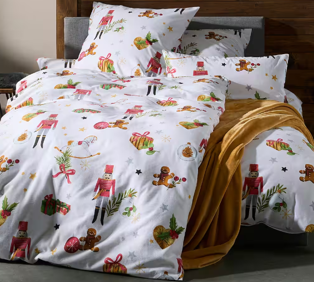 Bett mit weißer Winter-Bettwäsche, gemustert mit bunten Nussknackern, Lebkuchenmännern und Geschenken. Eine senfgelbe Decke liegt über dem Fußende, im Hintergrund ist ein Holzbett.