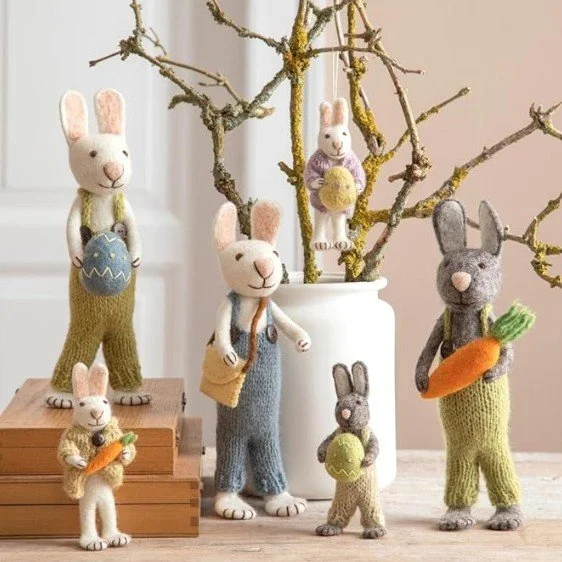 Gruppe handgefertigter Filz-Hasen mit Strickkleidung, die Ostereier und Karotten halten, dekoriert an Frühlingszweigen.