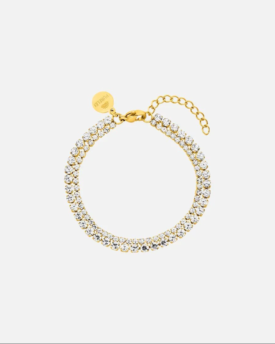 Goldenes, zweireihiges Tennisarmband von PURELEI mit rundum besetzten, klaren Glitzersteinen. Das Schmuckstück hat einen Karabinerverschluss, ein Verlängerungskettchen und einen Logo-Anhänger.