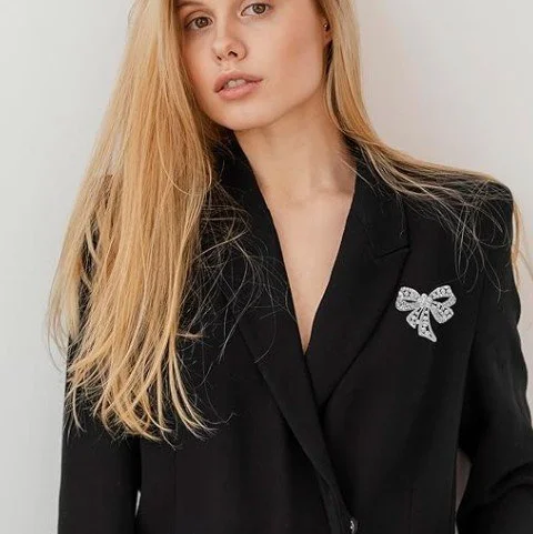  Nahaufnahme einer jungen Frau mit langen blonden Haaren, die einen schwarzen Blazer trägt. Am Revers des Blazers ist eine funkelnde, silberfarbene Brosche in Schleifenform mit Kristallbesatz befestigt. Heller, minimalistischer Hintergrund. 