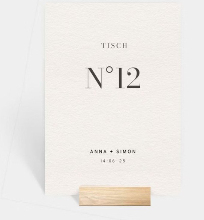  Eine schlichte, weiße Karte in einem Holzfuß mit der Aufschrift „TISCH N°12“ sowie den Namen „ANNA + SIMON“ und einem Datum. 