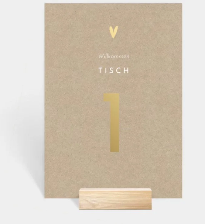  Eine moderne Karte aus braunem Kraftpapier mit einem kleinen goldenen Herz und der Aufschrift „Willkommen TISCH 1“ in Goldoptik. 