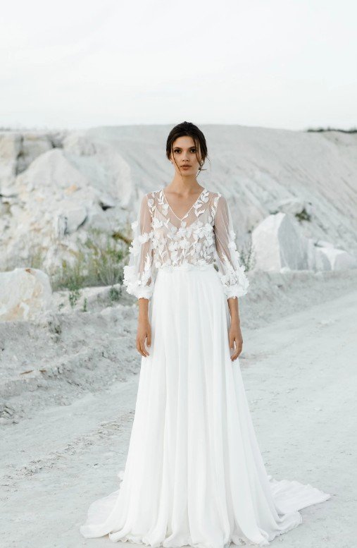 Ganzkörperaufnahme einer Braut in einer hellen Steinbruch-Landschaft. Sie trägt ein A-Linien-Kleid mit einem V-Ausschnitt und transparenten Ärmeln, die mit weißen Stoffblüten und Blattmotiven bestickt sind.