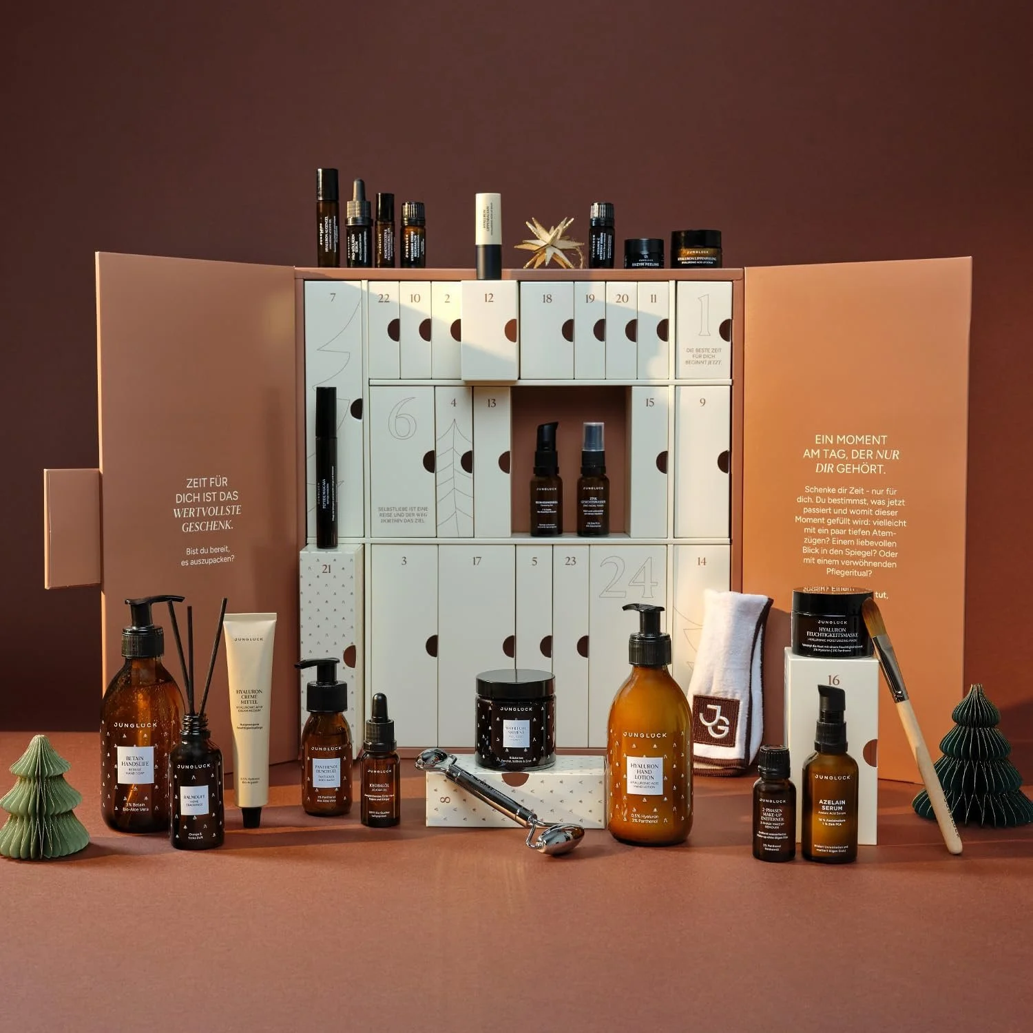 Ein geöffneter Junglück Beauty-Adventskalender in warmen Erdtönen mit hellen Schubladen. Um ihn herum sind die enthaltenen Pflegeprodukte in Braunglasflaschen und Tiegeln arrangiert.