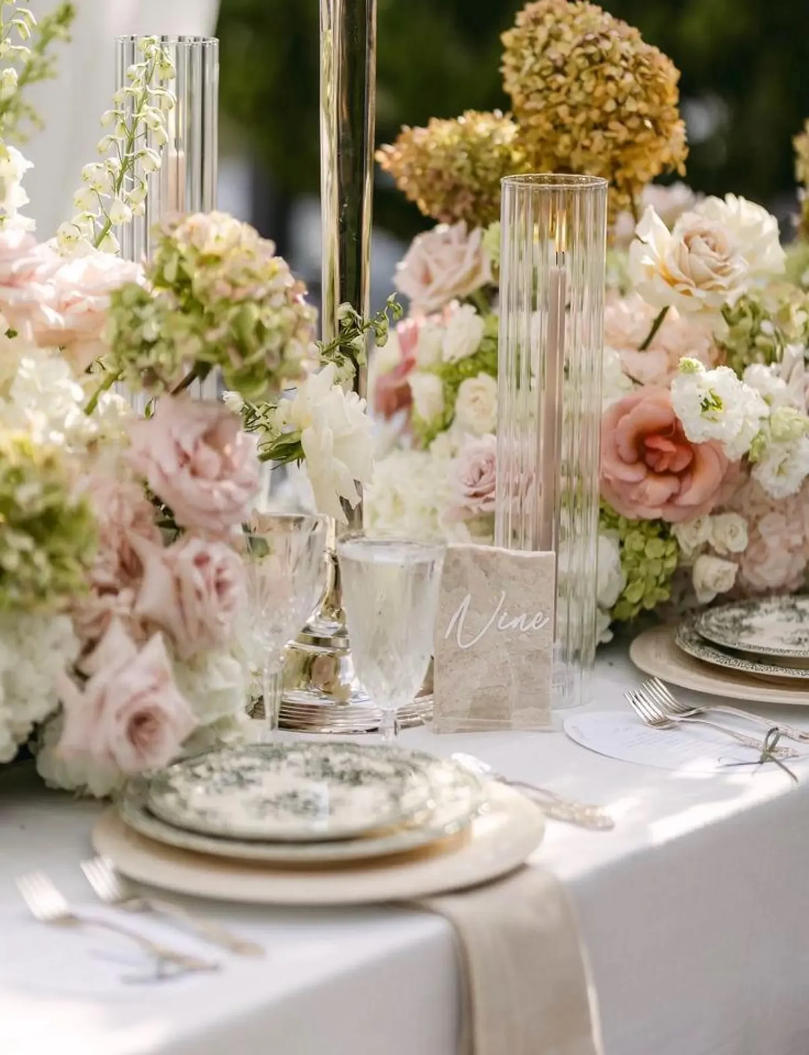 Ein elegant gedeckter Tisch im Freien, auf dem eine kleine, beige Marmorplatte mit der weißen Aufschrift „Nine“ dezent neben Blumengestecken platziert ist.