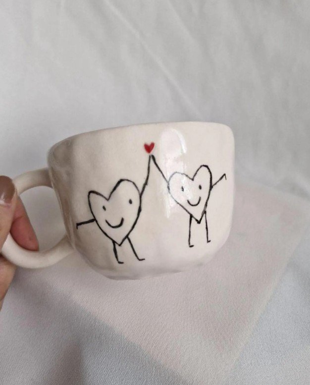 Handgefertigte Keramiktasse mit zwei gezeichneten Herzfiguren, die sich an den Händen halten und ein kleines rotes Herz zwischen sich hochhalten.