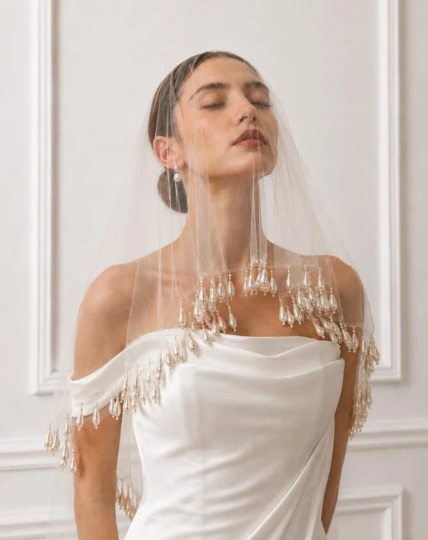 Braut mit geschlossenen Augen im schulterfreien Kleid und einem zarten Blusher-Schleier vor dem Gesicht, der mit hängenden Perlen-Tropfen besetzt ist. Inspiration für den Brautschleier-Guide.