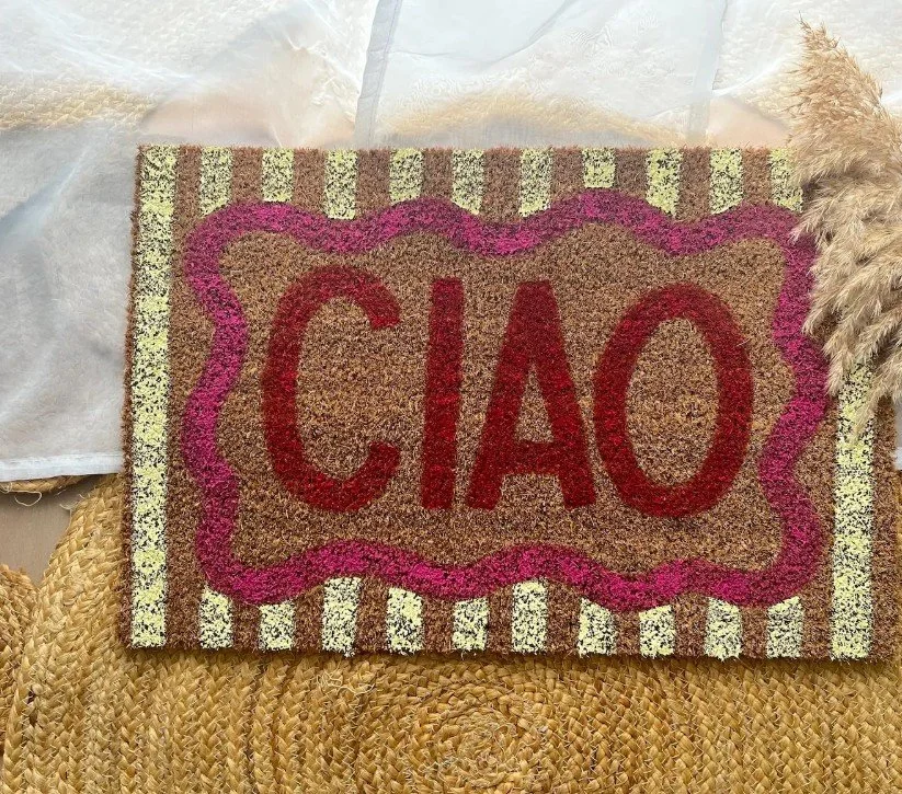Bunte Fußmatte aus Kokosfasern mit gelben Streifen, einem gewellten pinken Rahmen und dem großen roten Schriftzug „CIAO“ in der Mitte.