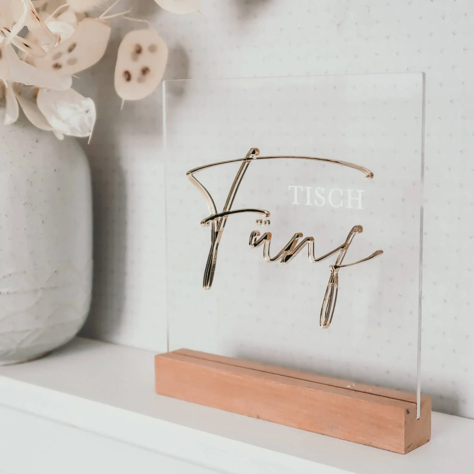 Eine moderne, quadratische Tischnummer aus klarem Acryl mit der eleganten goldenen Aufschrift „Tisch Fünf“ in einem hellen Holzfuß.