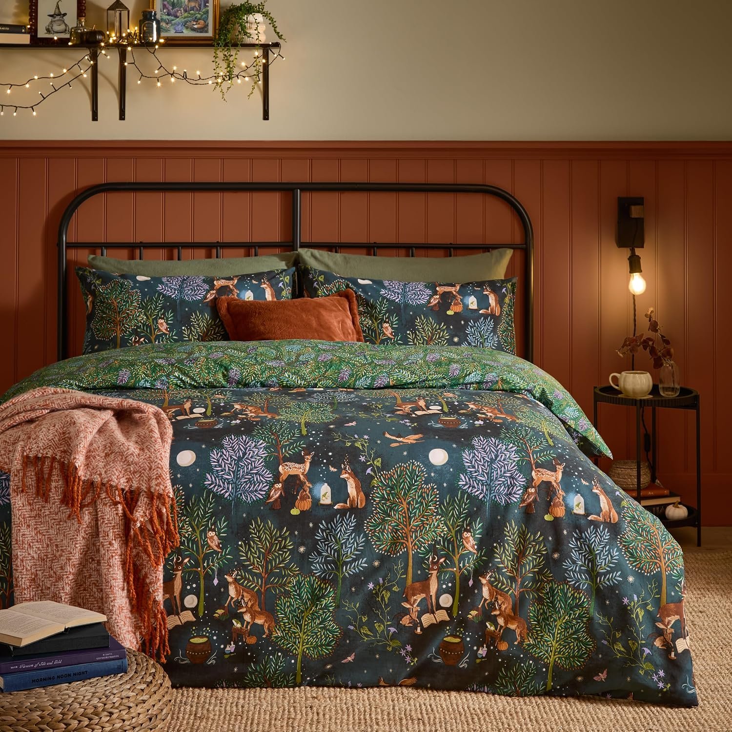 Dunkelblau-grüne Bettwäsche mit dichten Wald-, Fuchs- und Hirschmotiven auf einem Metallbett. Vor einer rötlichen Holzwand, dekoriert mit Lichterketten, Kissen und einer rötlichen Wolldecke.