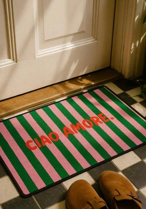 Moderne, gestreifte Fußmatte in Pink und Grün mit dem roten Text „CIAO AMORE.“ vor einer weißen Haustür auf kariertem Boden.