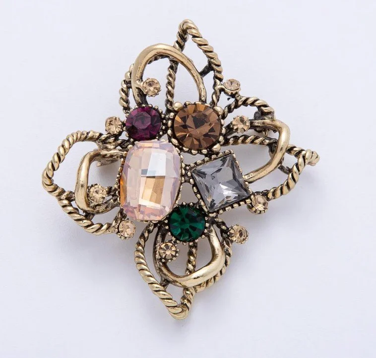 Goldene Vintage-Brosche mit Kordel‑Optik und Schmucksteinen: mittig rosafarbener und quadratischer grauer Stein, umgeben von runden Steinen in Grün, Violett und Braun. Vor weißem Hintergrund.