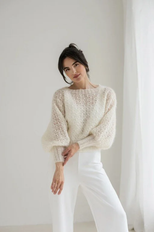 Brünette Frau trägt weißen, zart gemusterten Brautpullover mit leichtem Puffärmel und kombiniert ihn mit einer weißen, eleganten Hose in hellem Raum mit Tageslicht.